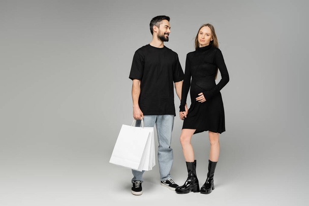 Joyful bearded man holding shopping bags and looking at trendy pregnant wife in black dress on grey background, new beginnings and parenthood, shopping and expectation concept - Fotografie, Obrázek