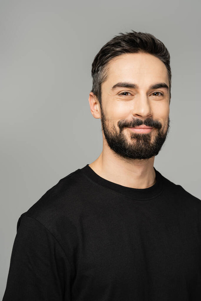 Portrait of positive bearded and brunette man in casual black t-shirt looking at camera while standing isolated on grey, masculine beauty concept, confident and charismatic  - Fotó, kép