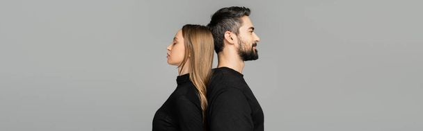 Side view of relaxed and bearded man in black t-shirt standing back to back with fair haired wife isolated on grey with copy space, husband and wife relationship concept, banner  - Photo, Image