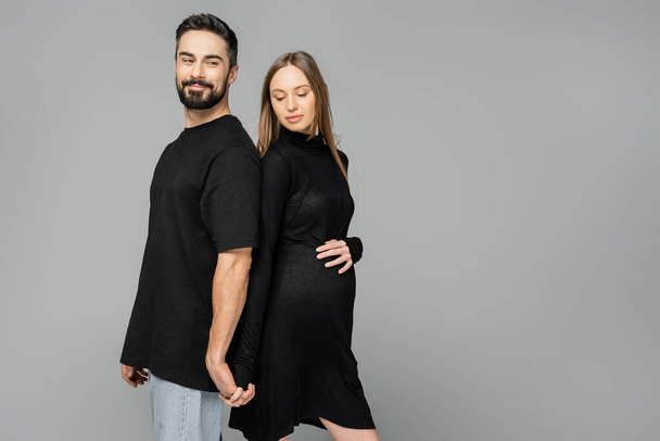 Positive bearded man holding hand of stylish pregnant wife in black dress and standing back to back isolated on grey, new beginnings and parenting concept   - Foto, imagen