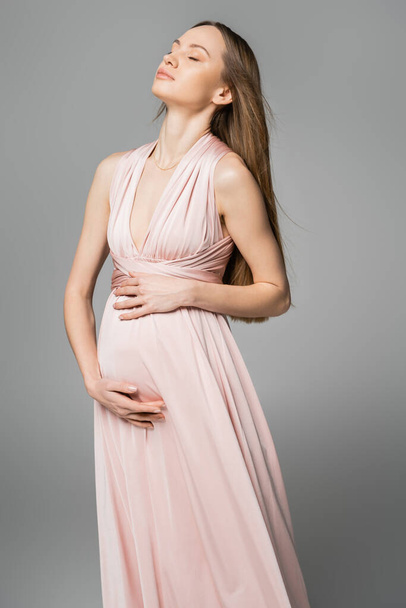 Entspannte hellhaarige und werdende Mutter im rosa Kleid, die den Bauch berührt, während sie posiert und isoliert auf grauer, eleganter und stilvoller Schwangerschaftskleidung steht, Sinnlichkeit, werdende Mutter  - Foto, Bild