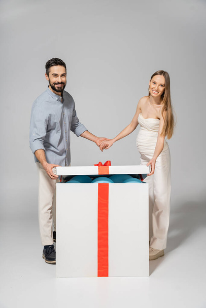 Cheerful and stylish couple holding hands while opening bog gift box with blue balloons during gender reveal surprise party on grey background, expecting parents concept, it`s a boy - Photo, Image