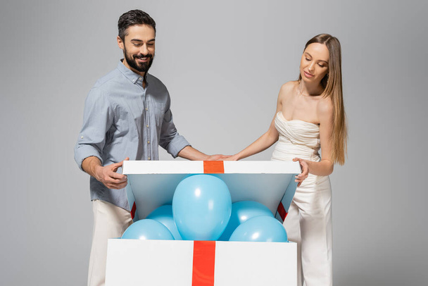 Cheerful and trendy expecting parents opening big gift box with festive blue balloons during gender reveal surprise party and celebration isolated on grey, it`s a boy - Foto, immagini