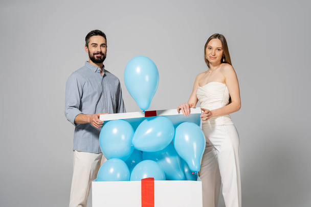 Positiv und stilvoll werdende Eltern blicken in die Kamera, während sie beim Öffnen einer großen Geschenkbox mit blauen Luftballons während des Geschlechtsverkehrs Überraschungsparty und Feier zeigen, isoliert auf grau, es ist ein Junge  - Foto, Bild