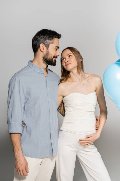 Smiling and stylish bearded man hugging and looking at pregnant wife while standing near blue festive balloons during gender reveal surprise party on grey background, expecting parents concept - Foto, afbeelding