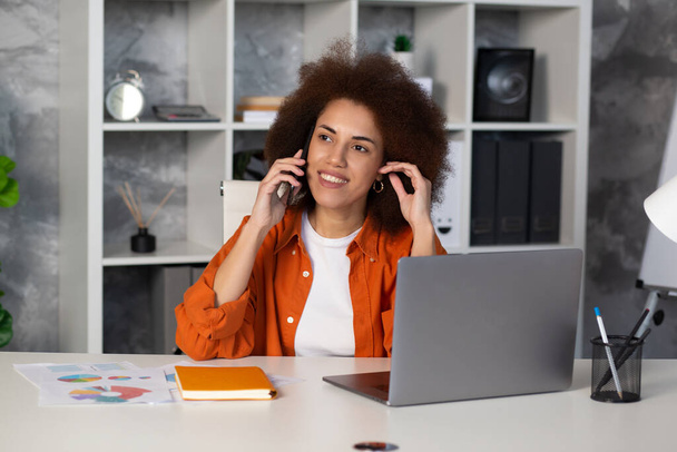 Succesvolle gelukkige Afro-Amerikaanse zakenvrouw glimlachend en pratend op een mobiele telefoon zittend op het werk in modern kantoor of coworking - Foto, afbeelding