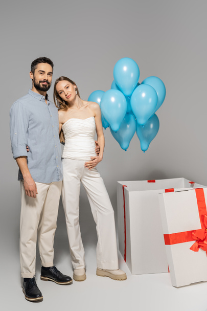 Voller positiver werdender Eltern, die sich umarmen und in die Kamera schauen, in der Nähe einer großen Geschenkbox mit festlich blauen Luftballons während der Geschlechterparty auf grauem Hintergrund, es ist ein Junge  - Foto, Bild