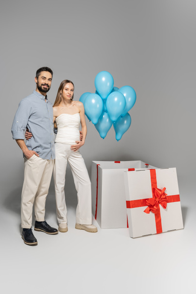 Full length of trendy and cheerful expecting parents hugging each other while standing near blue balloons and big gift box during gender reveal surprise party on grey background, it`s a boy  - Foto, imagen