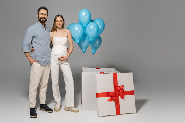 Pełna długość wesołych i modnych oczekiwań rodziców przytulających się i patrzących w kamerę stojąc w pobliżu dużego pudełka prezentów i świątecznych niebieskich balonów podczas płci ujawniają przyjęcie niespodzianka na szarym tle - Zdjęcie, obraz