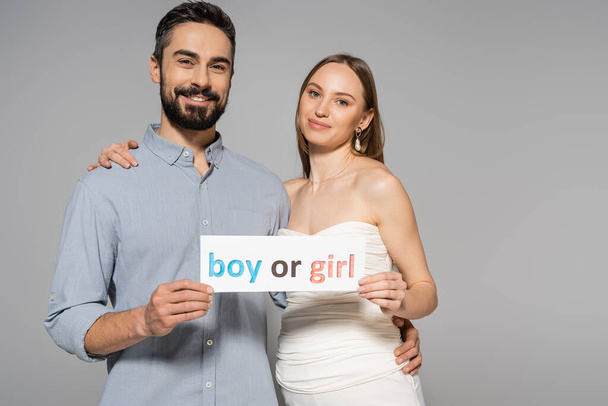 Positive and trendy pregnant woman holding card with boy or girl lettering and hugging stylish husband during gender reveal surprise party isolated on grey, expecting parents concept - Foto, Imagem