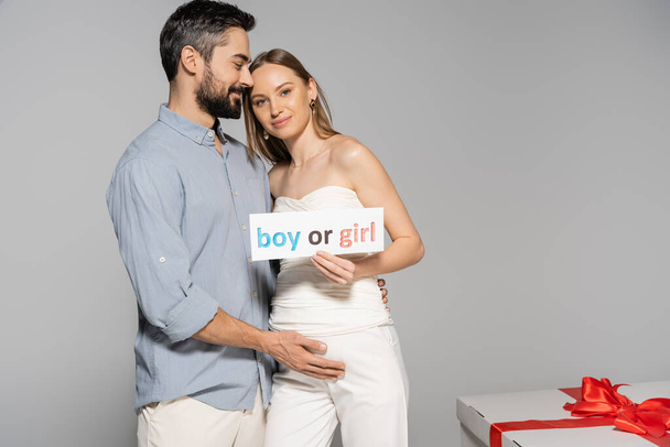Smiling pregnant woman holding card with boy or girl lettering near stylish husband and big gift box during gender reveal surprise party isolated on grey, expecting parents concept - Photo, Image
