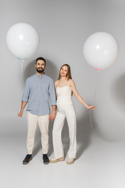 Volle Länge positiver und stilvoller werdender Eltern, die in die Kamera schauen, während sie Hände und Luftballons beim Geschlechtsverkehr halten, offenbaren Überraschungsparty auf grauem Hintergrund, erwartendes Eltern-Konzept - Foto, Bild