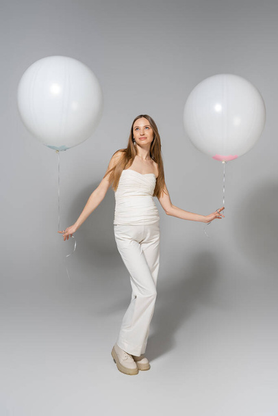 Full length of trendy and pregnant woman looking away while holding white festive balloons during celebration and gender reveal surprise party on grey background, fashionable pregnancy attire - Photo, Image