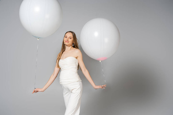 Mujer embarazada positiva y de moda mirando a la cámara mientras sostiene globos festivos blancos durante el género revelan fiesta sorpresa sobre fondo gris, atuendo de embarazo de moda - Foto, Imagen