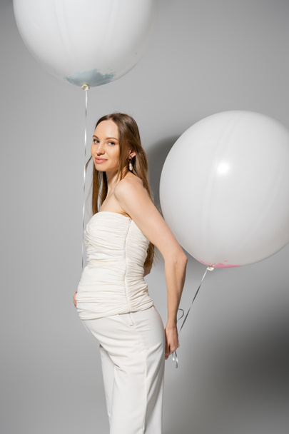 Χαμογελώντας και μοντέρνα έγκυος γυναίκα κοιτάζοντας κάμερα και κρατώντας λευκά εορταστικά μπαλόνια κατά τη διάρκεια του εορτασμού και το φύλο αποκαλύπτουν πάρτι έκπληξη σε γκρι φόντο, μοντέρνα ενδυμασία εγκυμοσύνης - Φωτογραφία, εικόνα