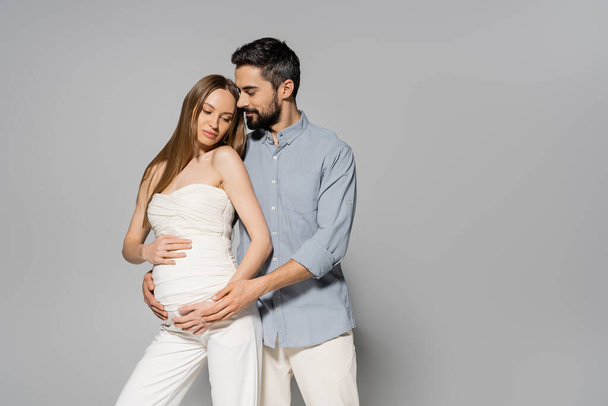 Trendy and bearded man hugging fair haired and pregnant woman while touching belly while standing together on grey background, expecting parents concept, baby bump, husband and wife  - Photo, Image