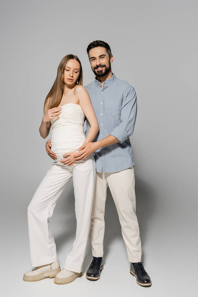 Full length of positive and bearded man hugging stylish and pregnant woman and looking at camera while standing together on grey background, expecting parents concept, baby bump, husband and wife  - Photo, Image