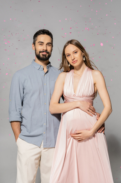 Portrait of smiling man hugging elegant and pregnant wife in pink dress while standing under confetti during baby shower party on grey background, expecting parents concept  - Foto, imagen