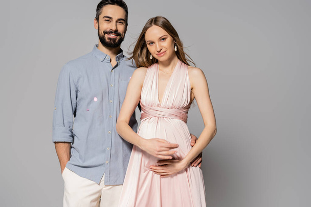 Stylish and cheerful man hugging elegant and pregnant wife while looking at camera and standing on grey background, expecting parents concept, new beginnings, parenthood  - Fotografie, Obrázek
