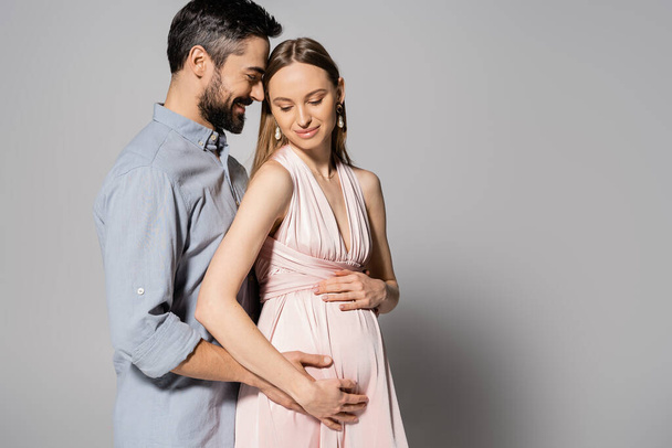 Happy and bearded man hugging pregnant woman in elegant dress while standing together on grey background, expecting parents concept, husband and wife  - 写真・画像