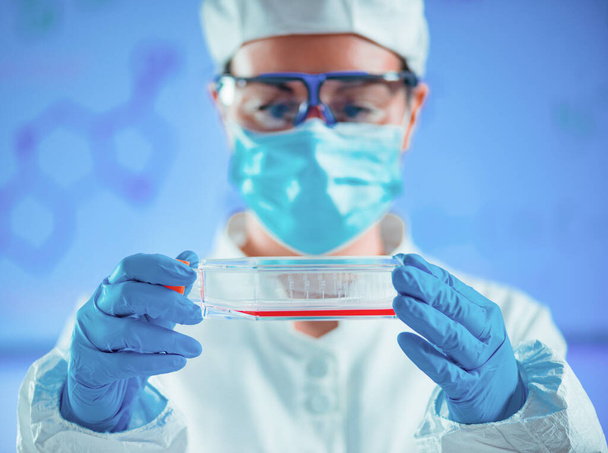 Laboratoriotakissa olevalla tiedemiehellä on pullo, joka on täytetty soluviljelmällä steriileissä olosuhteissa, mahdollisesti lääketieteellistä tutkimusta tai lääkekehitystä varten. Steriili ympäristö ja tarkat mittaukset ovat ratkaisevan tärkeitä kokeen onnistumisen kannalta.. - Valokuva, kuva