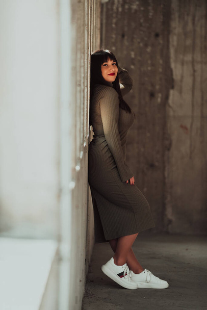 Eine selbstbewusste und stilvolle Plus-Size-Frau posiert vor einem modernen Gebäude in einem olivgrünen Kleid, das Schönheit, Ermächtigung und Selbstliebe ausstrahlt - Foto, Bild