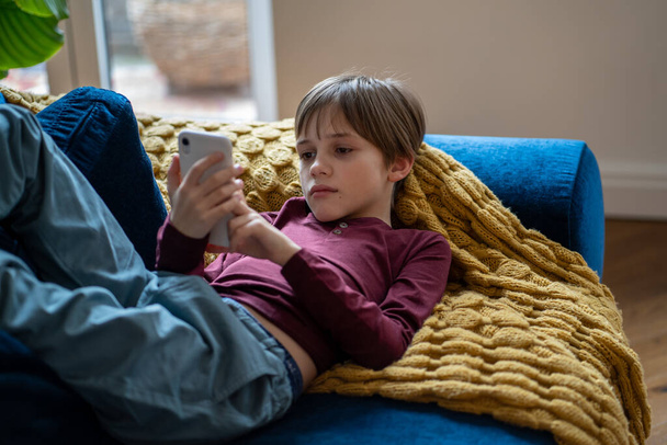 Ενδιαφερόμενο σχολιαρόπαιδο που χρησιμοποιεί smartphone. Περίεργο παιδί που κρατάει κινητό σε χέρια ξαπλωμένο σε καναπέ και κατεβάζει αιτήσεις στο σπίτι μόνο του. Χαλαρωμένο αγόρι που παίζει παιχνίδι κινητής συσκευής, βλέποντας κινούμενα σχέδια - Φωτογραφία, εικόνα