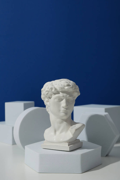 Геометрические фигуры и древняя голова на белом столе на синем фоне - Фото, изображение