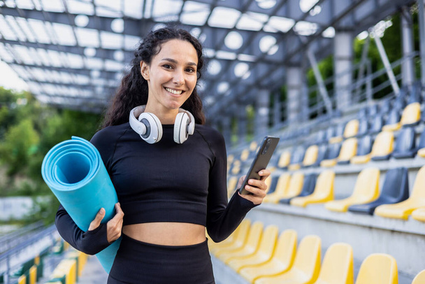 Portrait of happy female athlete, Hispanic woman standing near stadium outside smiling and looking at camera, holding phone and exercise mat, using app for co-training. - Foto, Imagen