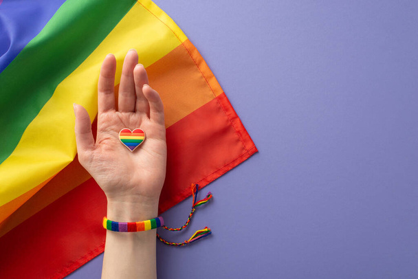 A first person top view of symbolic rainbow-colored bracelet adorns a girl's hand holding a heart-shaped badge over flag on a lilac background with space for text, recognizing LGBTQ History Month - Fotó, kép