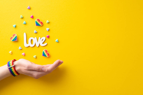 In this image, woman's hand donning a symbolic wristlet with pin badges, inscription love and hearts flying out from her palm takes center stage against yellow backdrop with space for text or ad - Фото, изображение