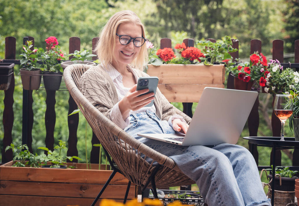 Beautiful blond middle-aged woman relaxing on the terrace of a country house in summer and using a laptop for online work - Фото, изображение