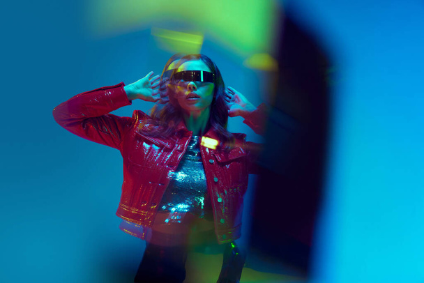 Porträt mit attraktivem Model in trendiger Kleidung mit roter Lederjacke und Sonnenbrille über blauem Neon-Hintergrund. Glitch-Effekt. Konzept der Schönheit, Fashion Week, Emotionen, Trends, Werbung - Foto, Bild