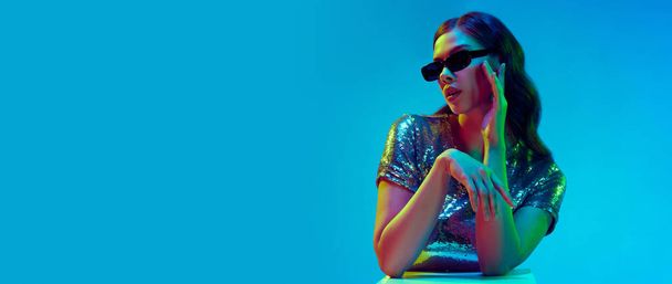 Banner with attractive fashion model, dreaming girl wearing trendy clothes, silver top and black sunglasses over blue background in neon light. Concept of beauty, fashion week, human emotions, ad - Foto, afbeelding