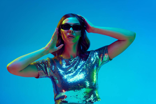Beautiful fashion model, dreaming woman wearing fashionable clothes, silver top and black sunglasses posing over blue background in neon light. Concept of beauty, fashion week, human emotions, ad - Photo, Image