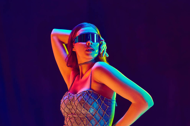 High fashion model dragen futuristische stijl van kleding met zwarte zonnebril poseren over violette studio achtergrond in neon licht. Sluit maar af. Schoonheidsbegrip, modeweek, menselijke emoties, trends, reclame - Foto, afbeelding