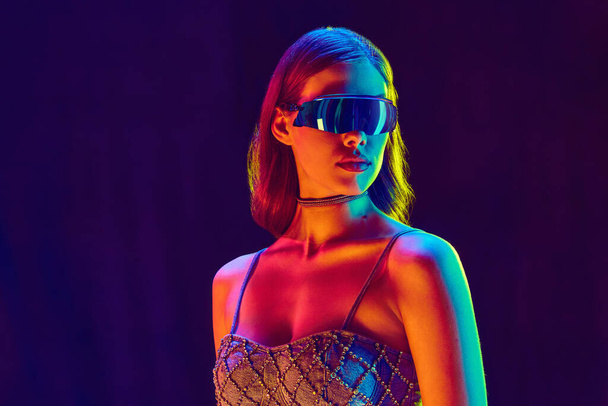 Beautiful fashion model, serious woman wearing fashionable clothes, crop top and black sunglasses posing over violet background in neon light. Concept of beauty, fashion week, human emotions, ad - Photo, Image