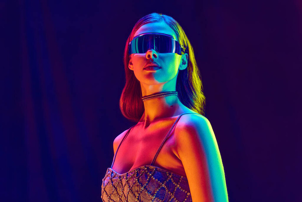 Modelmodel, junges Mädchen mit langen Haaren und trendiger Sonnenbrille in einem schwarz glänzenden Oberteil, das vor dunklem Studiohintergrund in Neonlicht posiert. Schönheitskonzept, Modewoche, menschliche Emotionen, Werbung - Foto, Bild