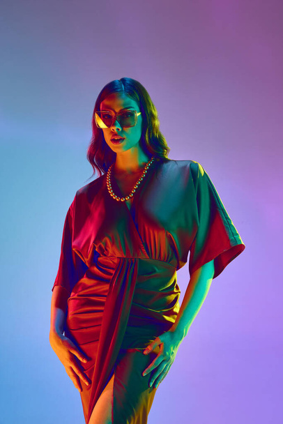 Attraktive Frau, Modemodel, trägt elegantes Seidenkleid mit Halsketten und trendige Sonnenbrille über violettem Hintergrund in Neonlicht. Schönheitskonzept, Modewoche, menschliche Emotionen, Werbung - Foto, Bild