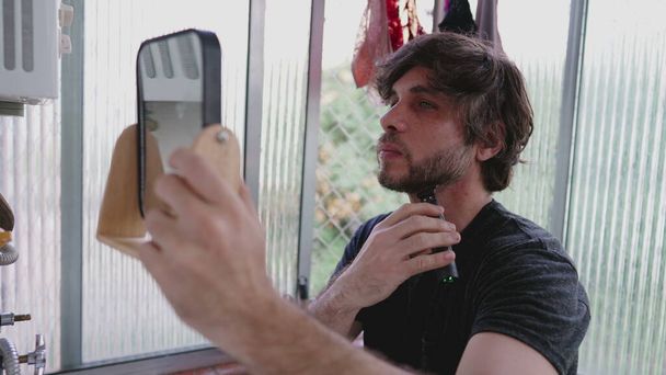 One young man shaving beard with trimmer on the go while standing in apartment balcony and holding a mirror - Foto, afbeelding
