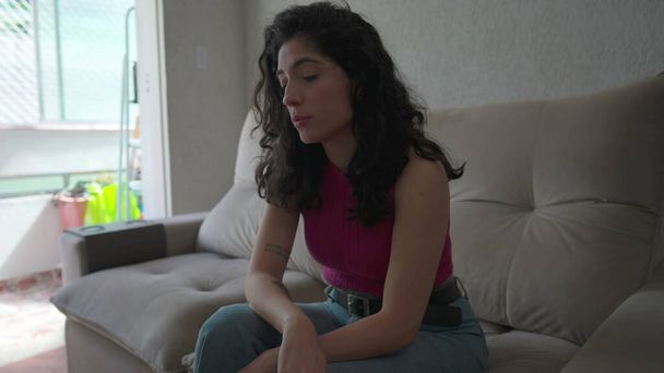 Una joven pensativa sentada en el sofá de su casa con expresión contemplativa. Persona femenina de 30 años en dilema - Foto, imagen