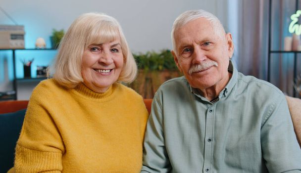Glückliches älteres älteres Familienpaar, das sich umarmt, lacht, lächelnd in die Kamera schaut, gesunde Rentner mit zwei Großeltern. Großmutter Großvater umarmt sitzen auf dem Sofa zu Hause, Porträt - Foto, Bild