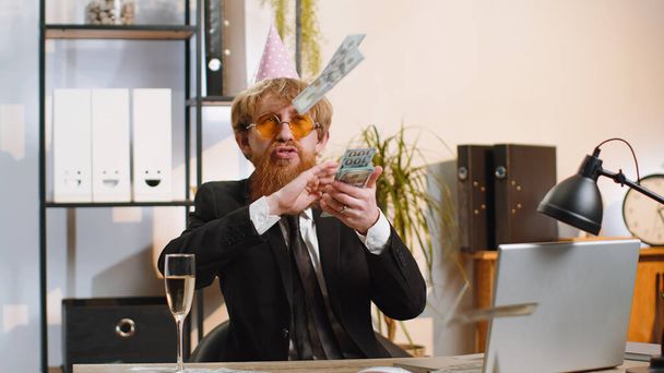 Cheerful rich young businessman working on laptop and wasting throwing money to camera more tips earnings big profit success win lottery shopping. Happy freelancer bearded man at home office desk - Foto, Imagen