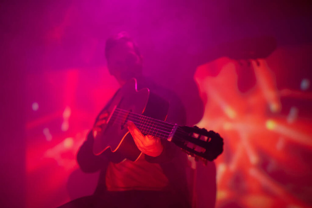 Músico tocando guitarra acústica em um clube nebuloso com luzes coloridas. - Foto, Imagem