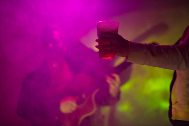 Woman holding a plastic beer cup with a musician playing guitar in the club background. - Photo, Image
