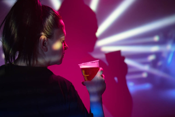 Woman holding and drinking a plastic cup of beer in the nightclub. - Photo, Image