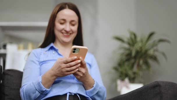 Gelukkige casual mooie vrouw praten aan de telefoon terwijl zitten op de bank thuis. glimlachende jonge vrouw met behulp van mobiele telefoon thuis. - Video