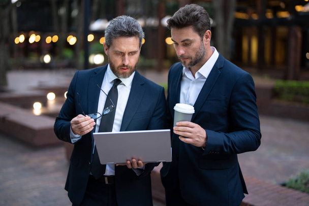Two handsome young businessmen in classic suits using laptop. Business men team using laptop outdoor. Businessmen looking laptop with their business success in city. Two business teams success - Fotografie, Obrázek