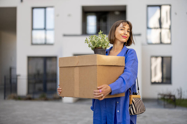 Jonge vrouw met kartonnen doos en bloempot op de binnenplaats van het appartementencomplex, verhuizen naar een nieuw huis - Foto, afbeelding