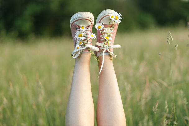 Woman legs in pink sneakers with daisy flowers in summer countryside, close up. Young female relaxing among wildflowers in meadow. Carefree and summer vacation, funny moment - Фото, изображение